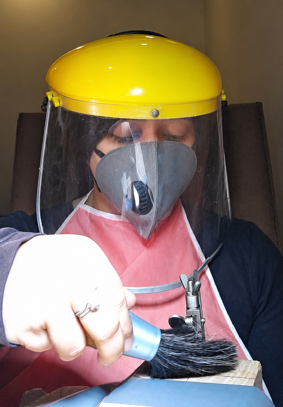 Maadin Artistry artisan in a yellow face shield, respirator mask and pink apron polishing a ring at the workbench